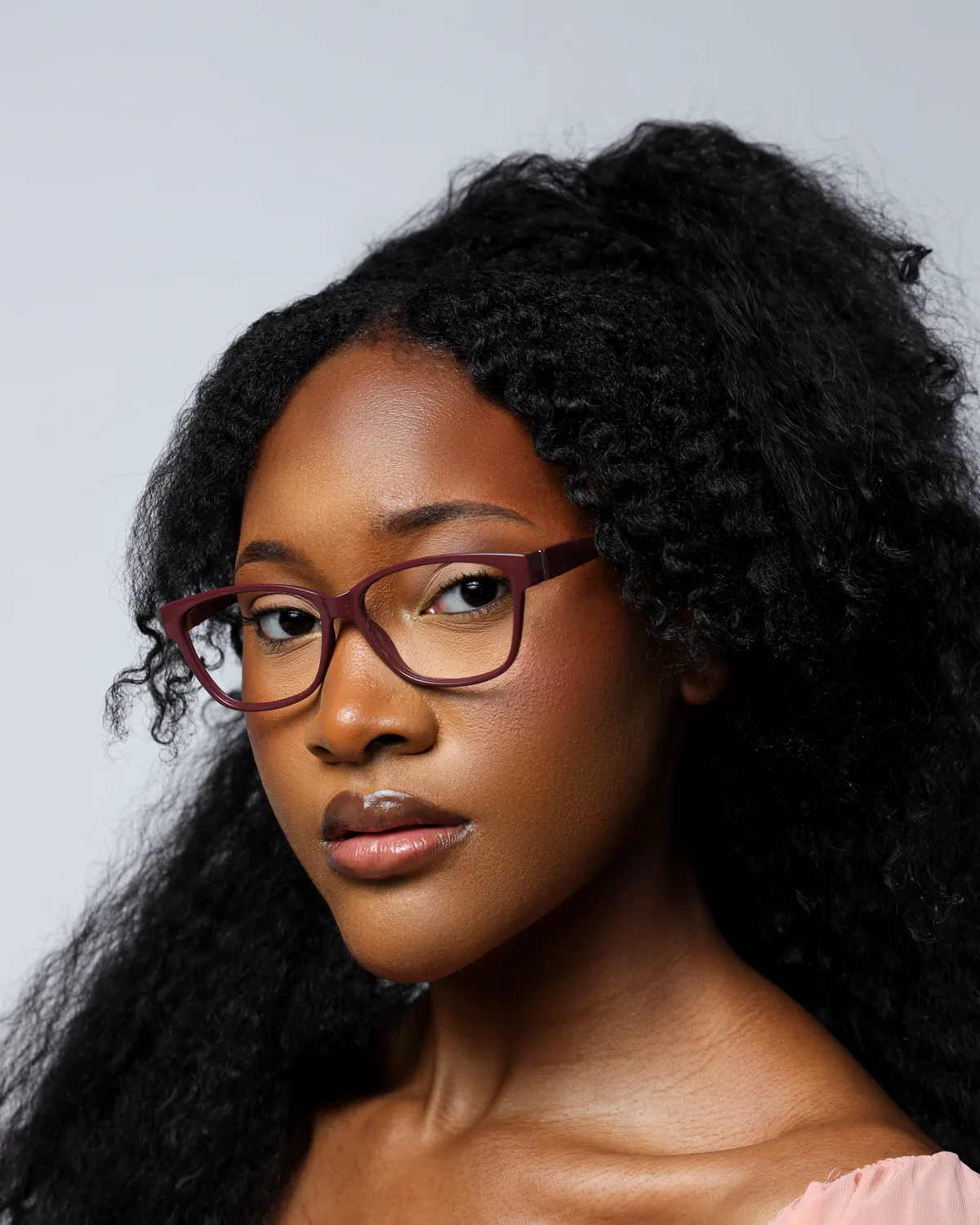 Ahdis Beruk with her hair down, in plum glasses and a peach top, against a grey background
